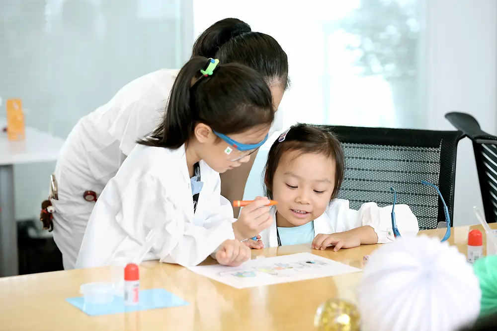 Laboratuvar önlüğü giymiş bir kadın ve iki çocuk bir masada resim yapıyor