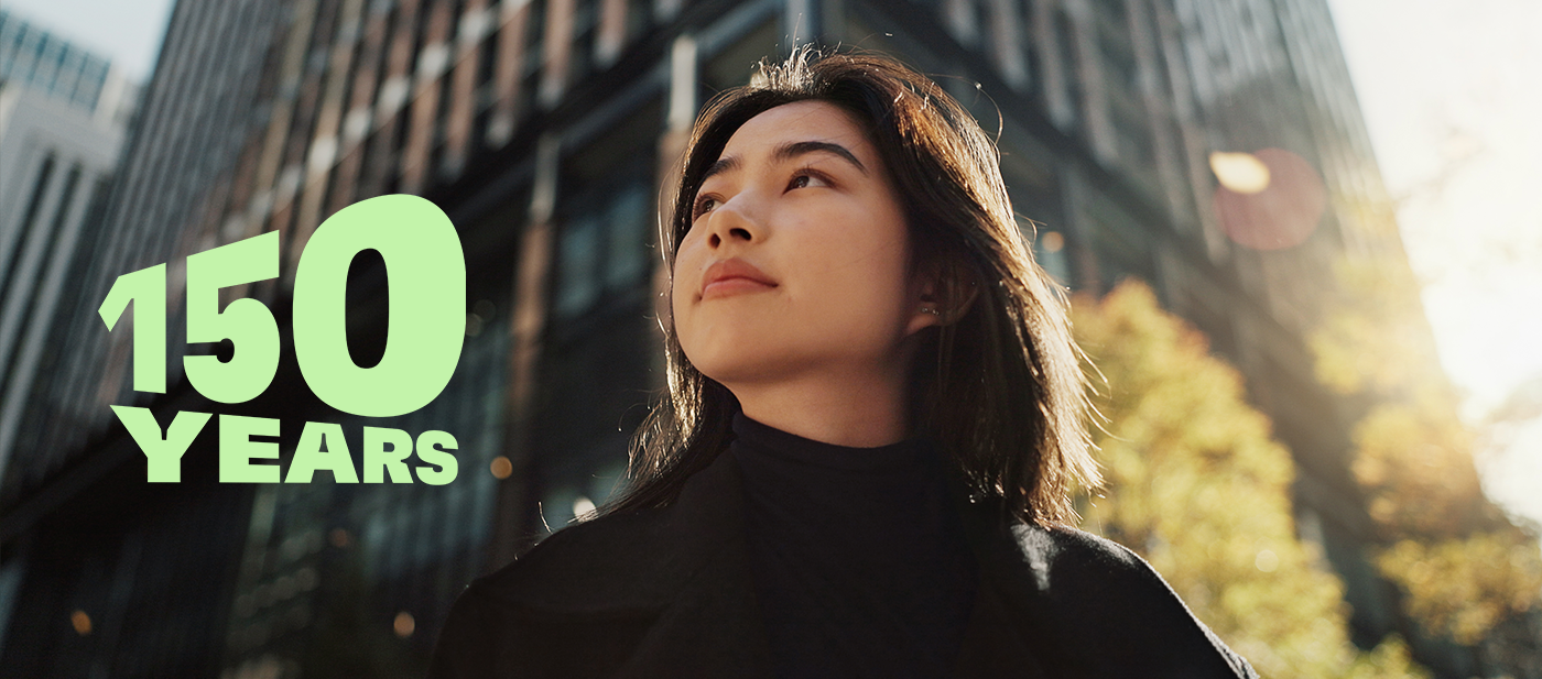 Yüksek cam binalar ve sonbahar ağaçlarıyla modern bir şehir manzarası üzerinde yer alan yeşil “150 YEARS” yazısı, 150. yıl dönümü kutlamasını simgeliyor.