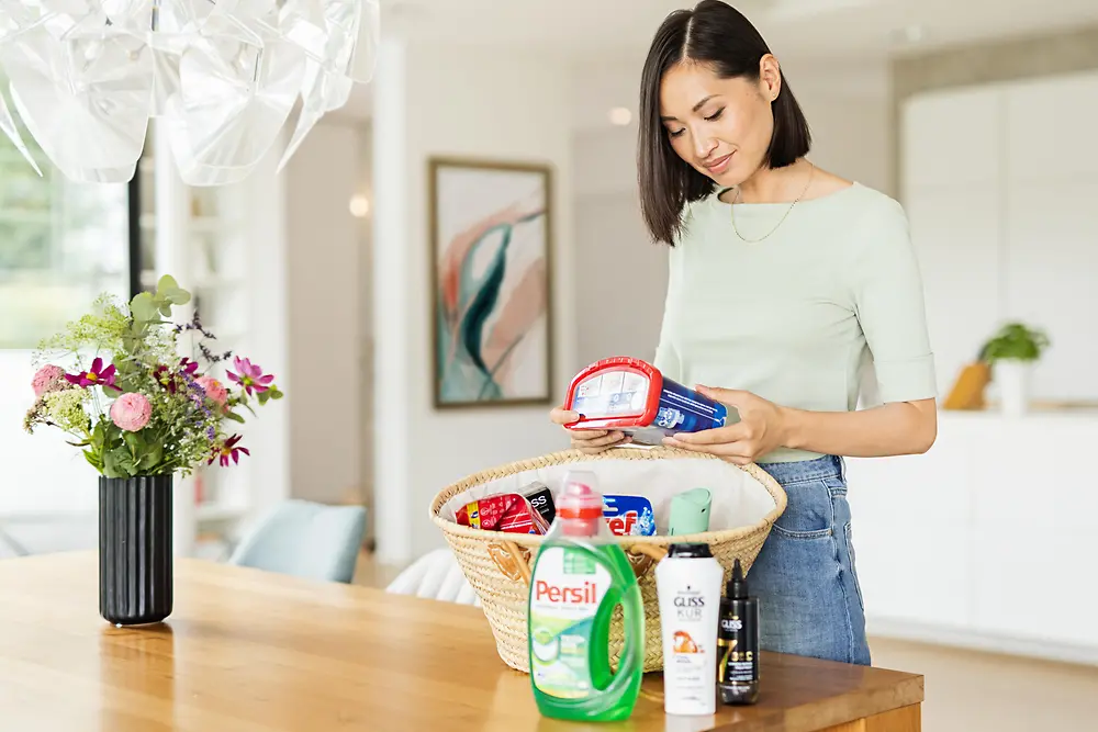 Bir kadın; Persil, Gliss, Bref, Somat, Taft gibi Henkel ürünleriyle dolu bir sepetin durduğu masanın yanında.