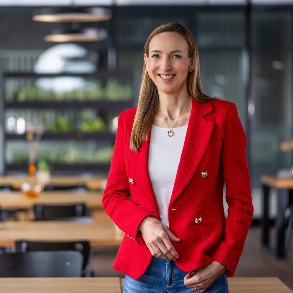 Modern, kafe tarzı bir ortamda; ahşap masalar ve dekoratif vazolar eşliğinde, beyaz bir üst ve parlak kırmızı bir ceket giymiş Dr. Simone Bagel-Trah.