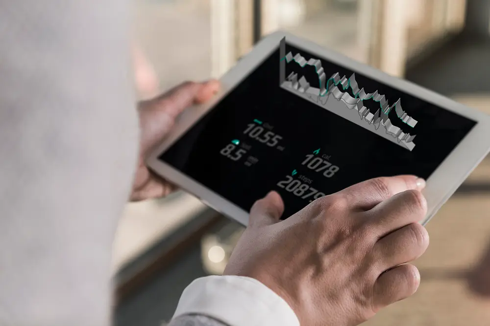 Aydınlık bir iç mekânda, omuz üzerinden görülen bir açıyla, dijital grafikler ve sayısal veriler görüntülenen bir tablet tutan kişi.