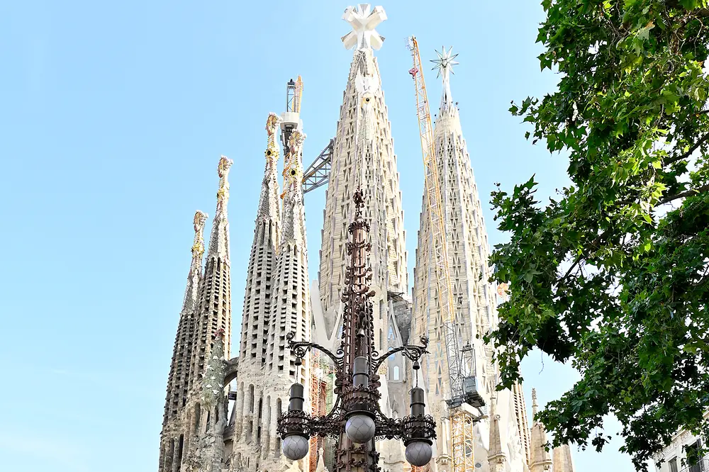 Vista general de la Sagrada Familia en Barcelona con sus torres centrales en construcción y cuatro personas posando en primer plano frente al templo en una plaza urbana con peatones y elementos de mobiliario urbano.